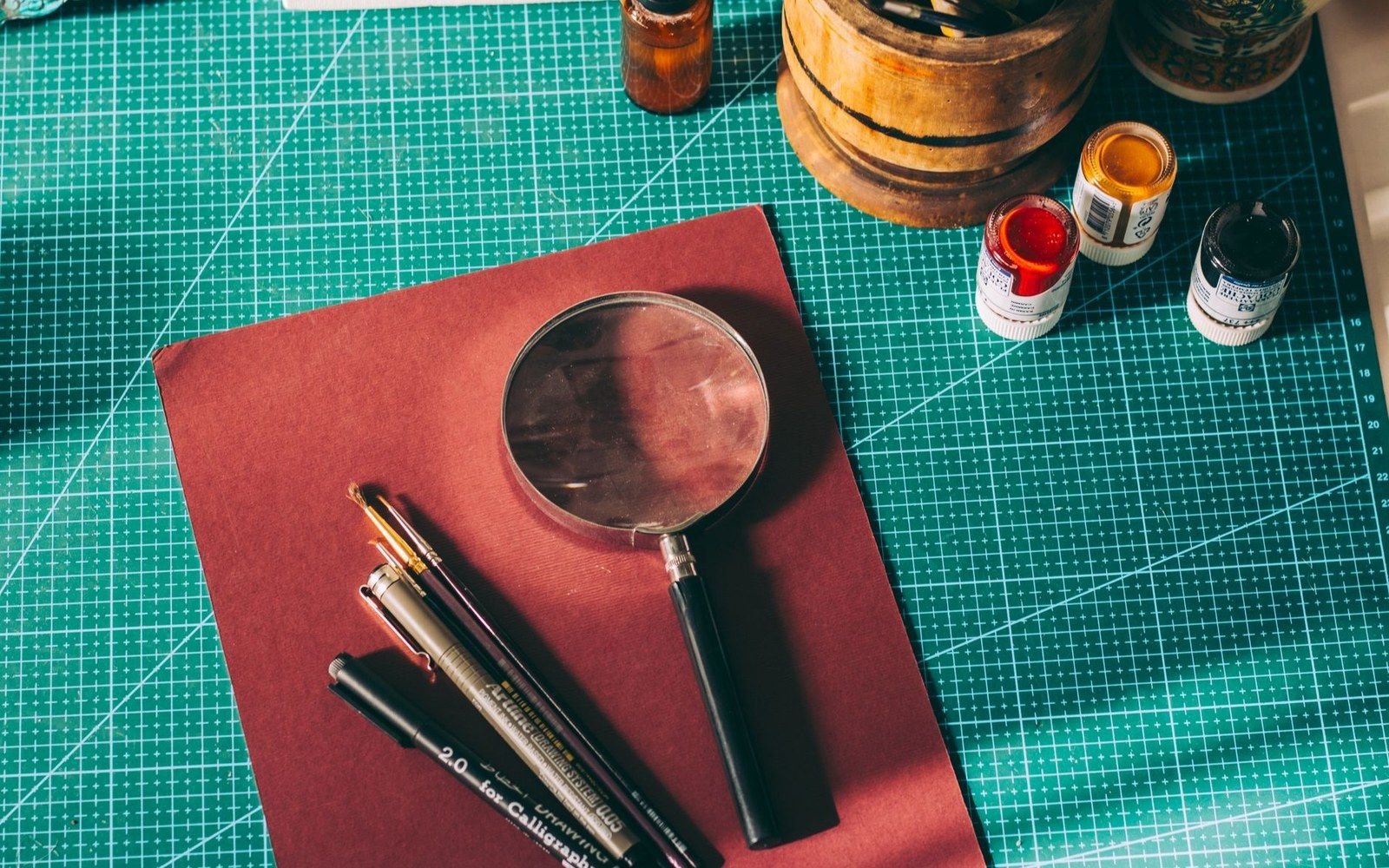 Magnifying glass, pens and small paint jars arranged on a cutting mat