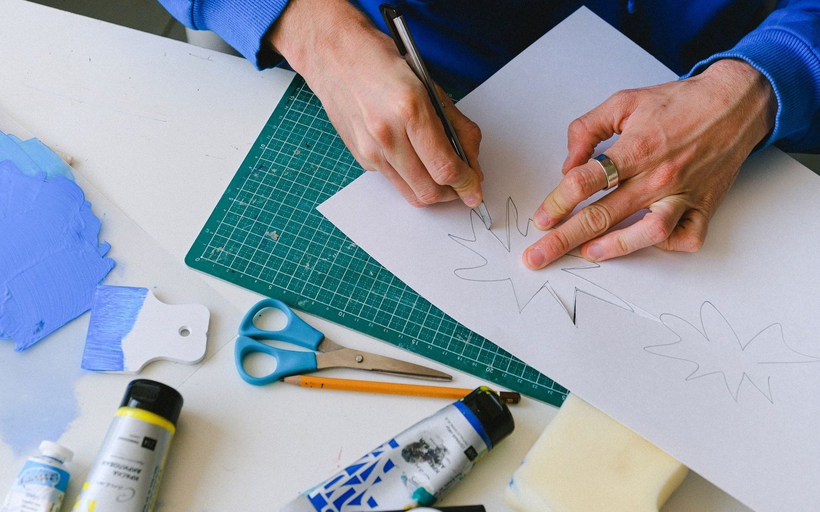 Hands sketching a leaf-shaped stencil on paper beside scissors and paint tubes